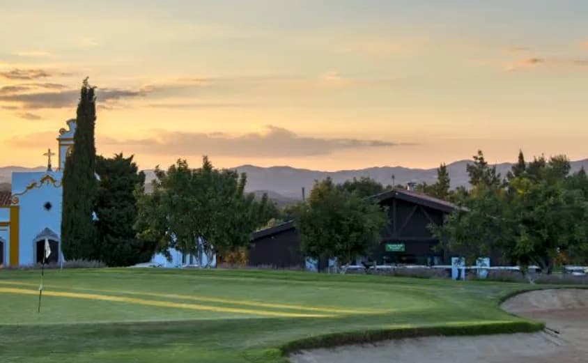 Green at Benamor Golf at sunset, beside a small blue-and-yellow chapel, cypress trees and distant hills